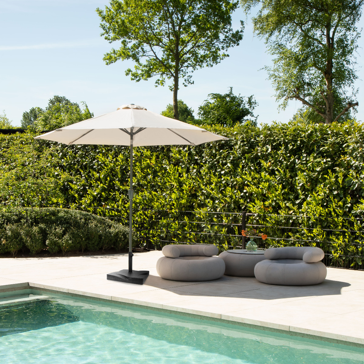 A patio umbrella stands by a pool with seating and a hedge.