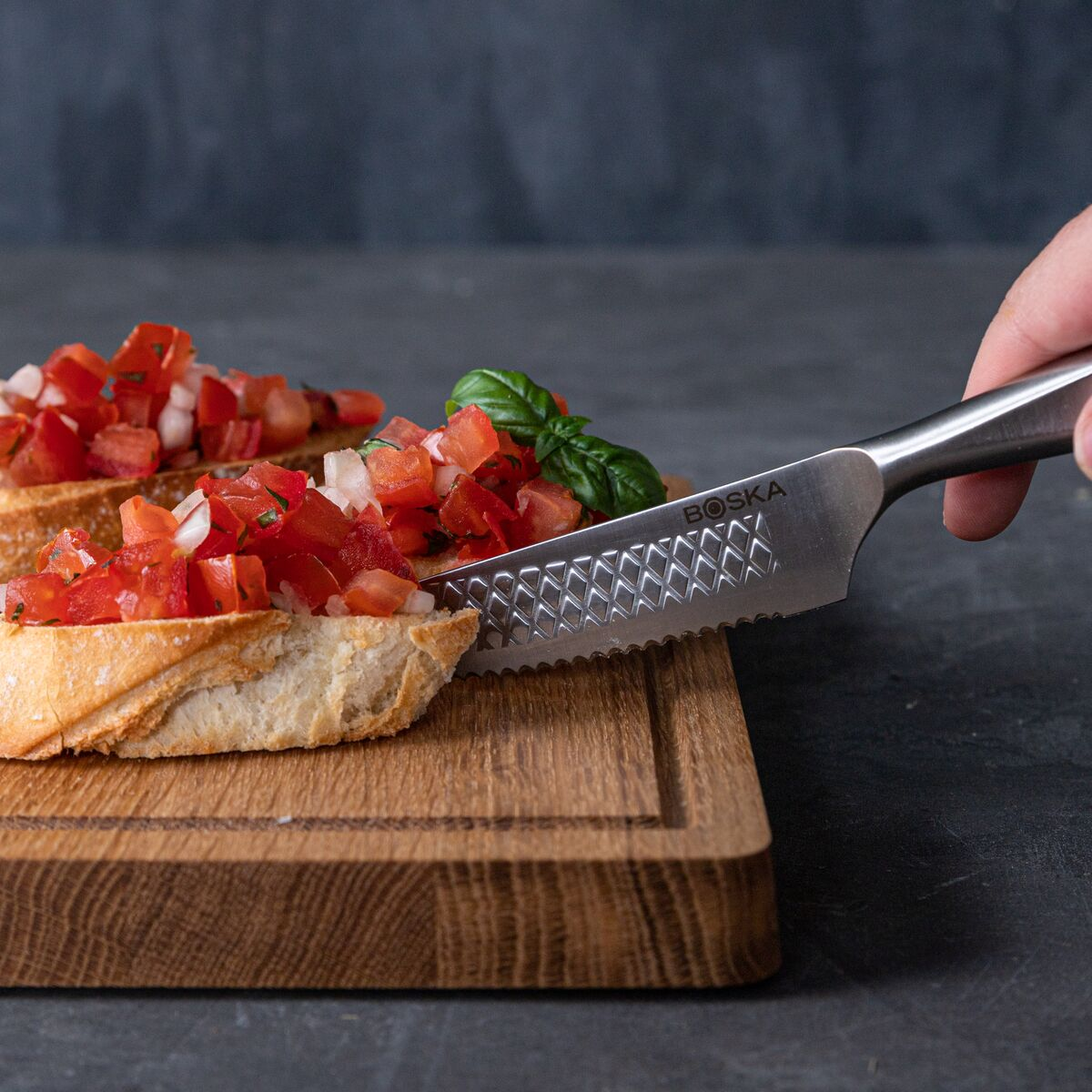 A knife cuts bruschetta with tomatoes and basil on a wooden board.