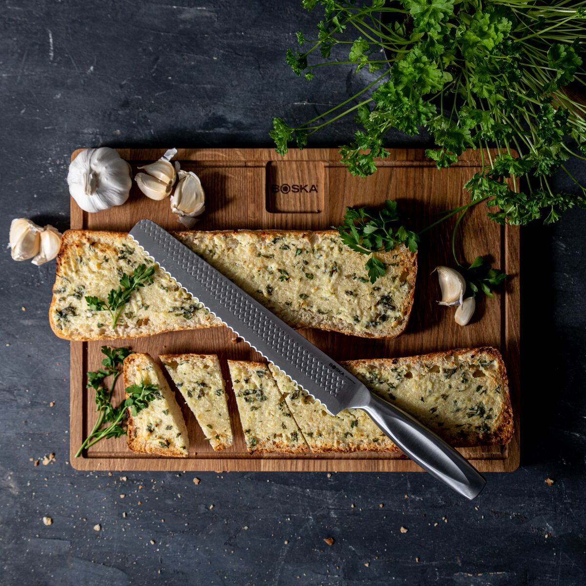 Garlic bread with parsley on a wooden board, next to garlic and a knife.
