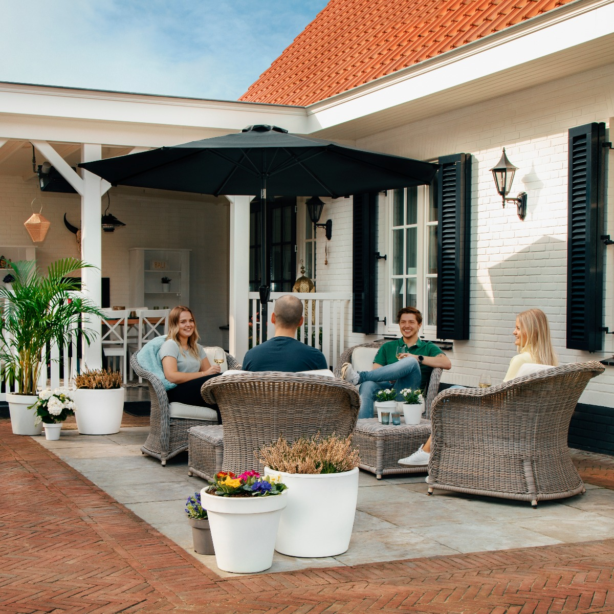 Vier mensen ontspannen op een terras buiten op rieten meubels onder een parasol.