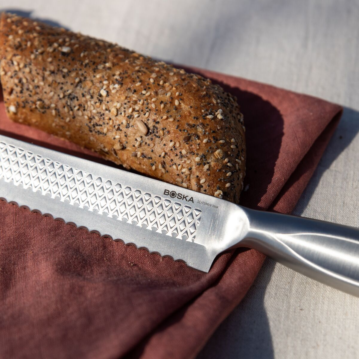 A bread knife lies next to a seeded roll on a red cloth.