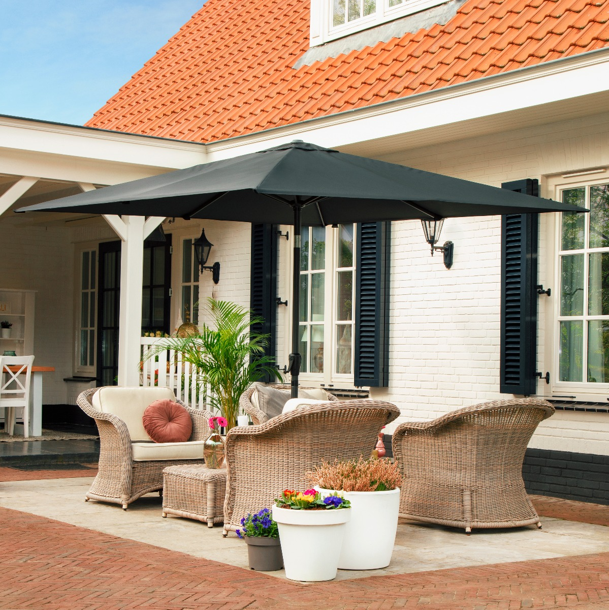 Terras met rieten banken, parasol en bloemen voor een wit huis.