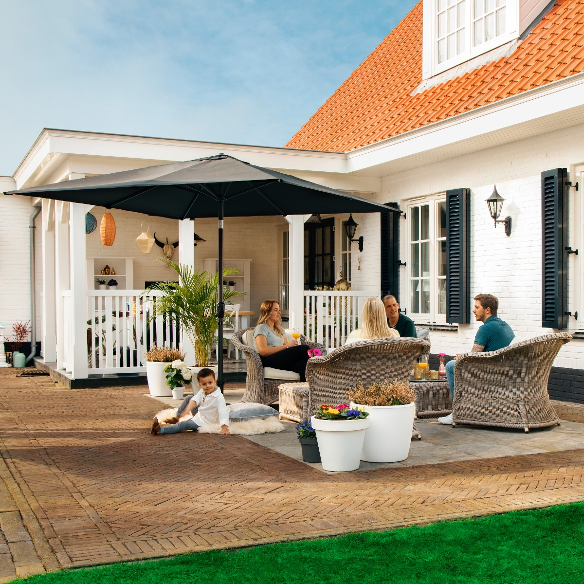 Familie ontspant op een terras onder een parasol, een kind speelt op de grond.