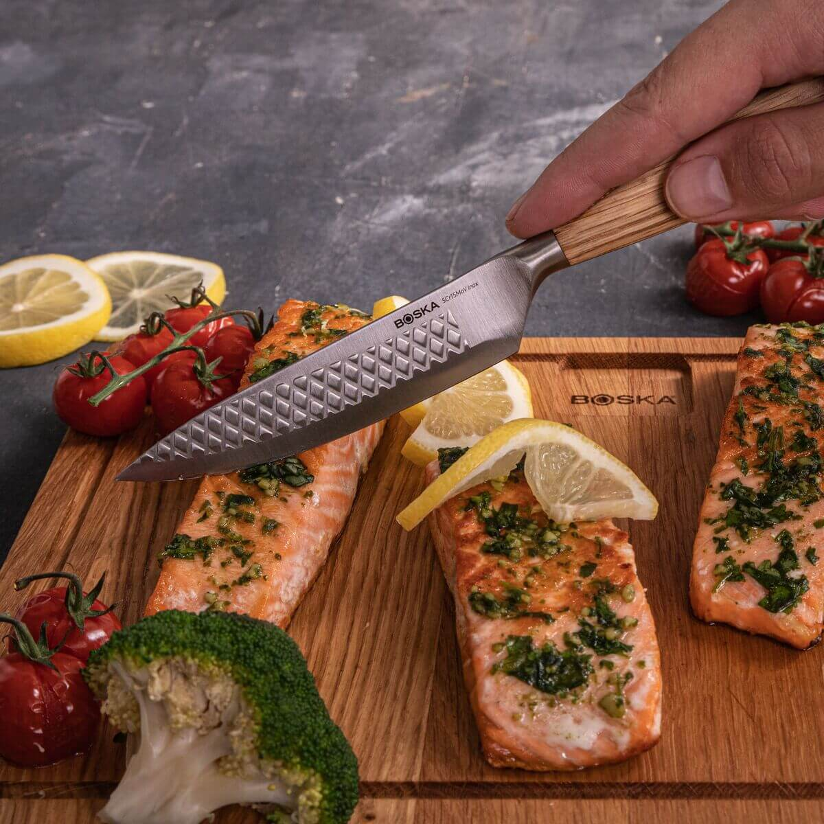 A knife over grilled salmon on a wooden board with tomatoes and lemon.