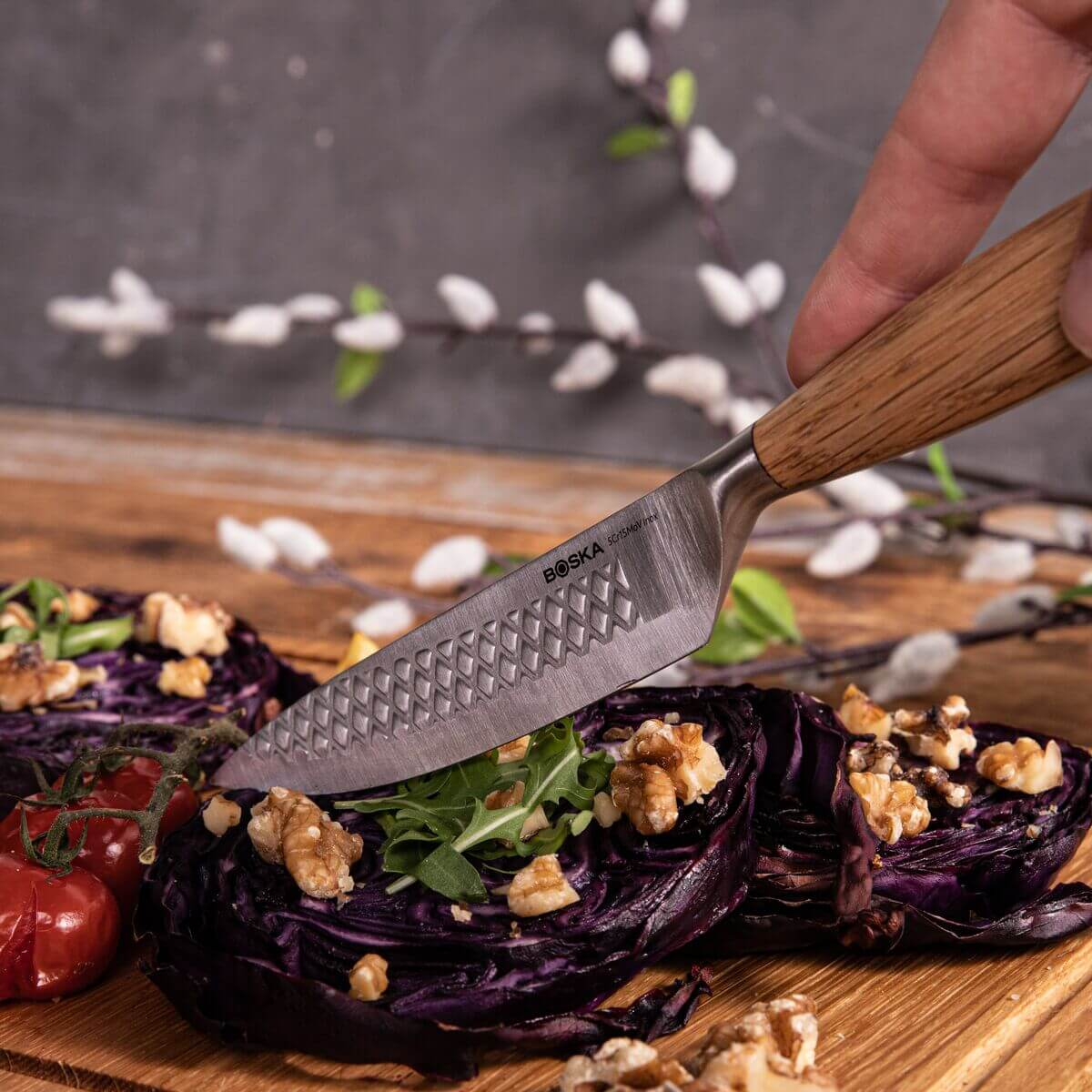 A knife with a wooden handle cuts red cabbage on a wooden board, garnished with walnuts and tomatoes.