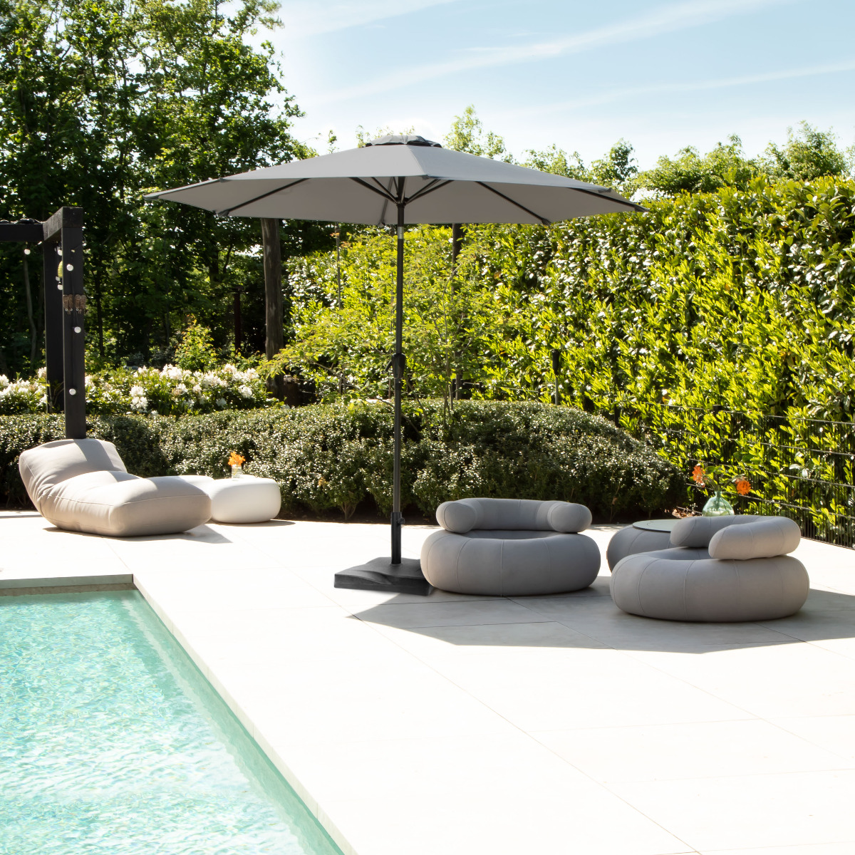 Outdoor furniture by pool: gray chairs, table, umbrella, green hedge.
