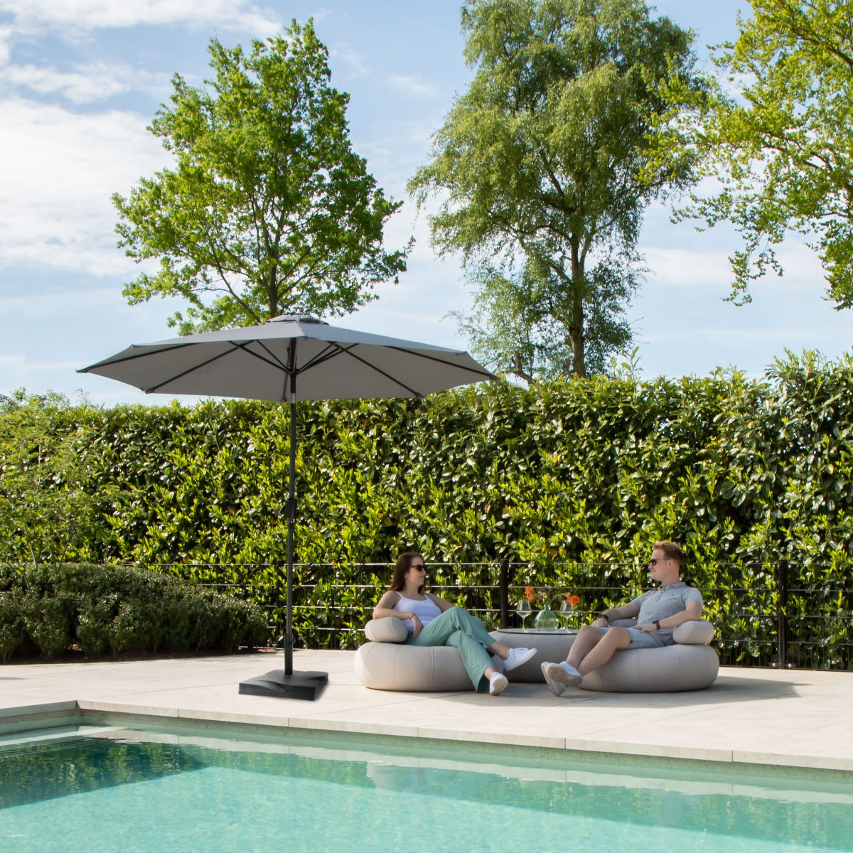 A couple relaxing on a sofa by the pool under an umbrella, surrounded by greenery.