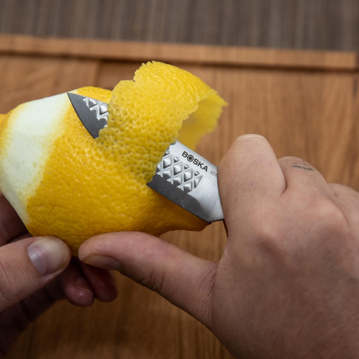 Hands peeling a lemon with a zester on a wooden board.