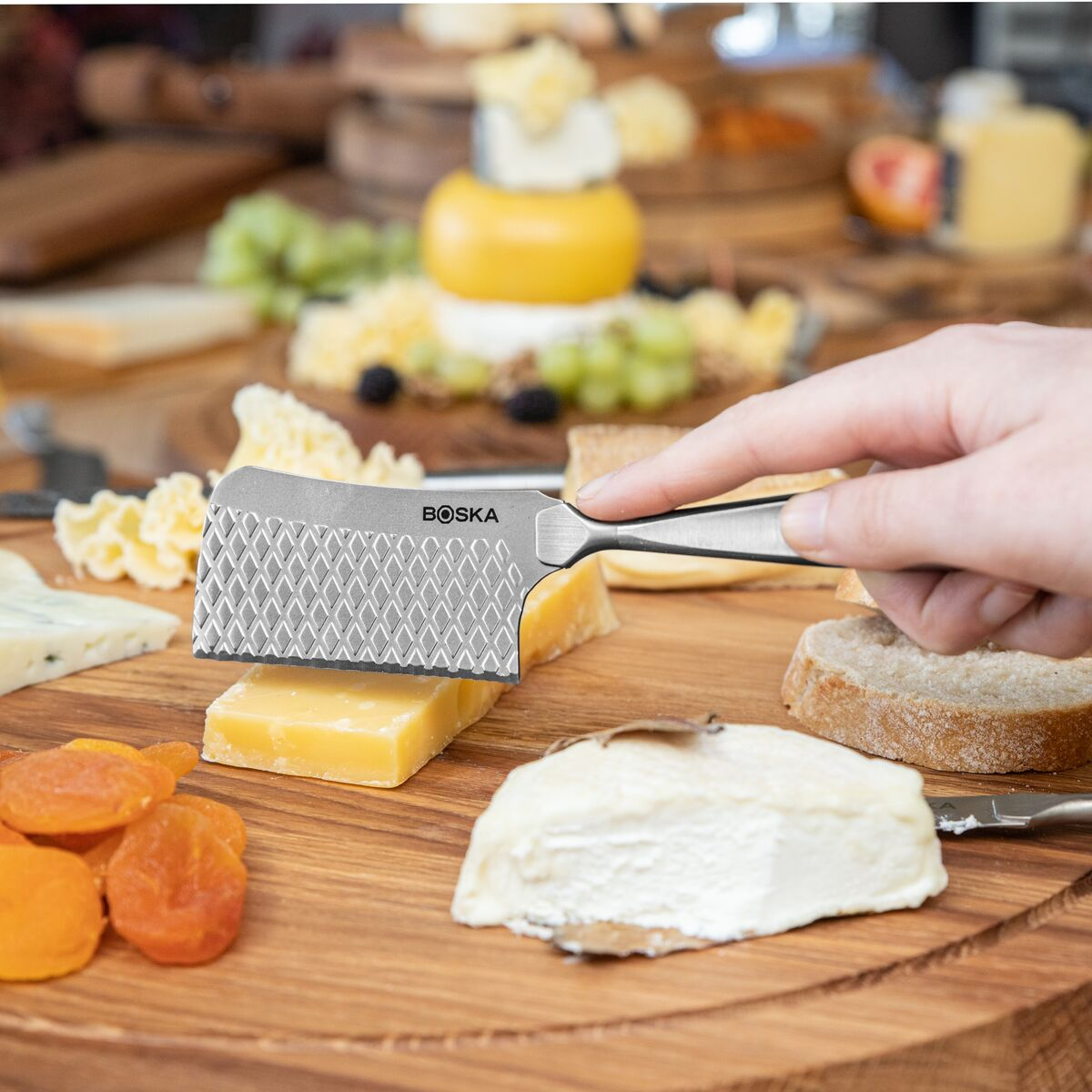 A hand holds a BOSKA cheese knife over a cheese board with various cheeses, fruit, and bread.