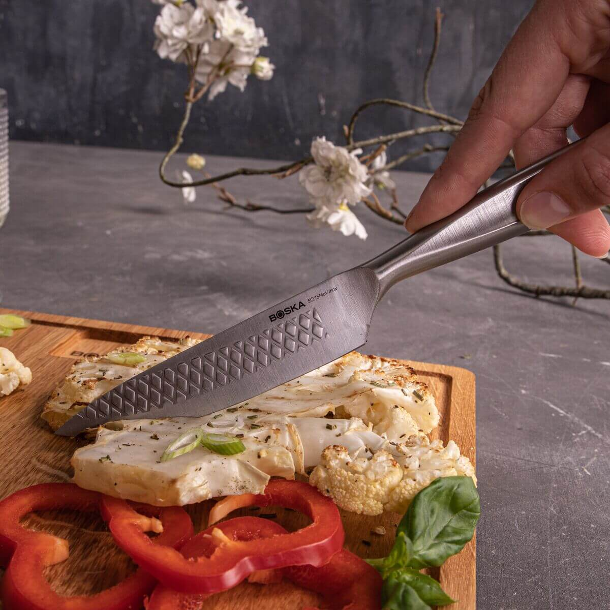 A knife cuts grilled cauliflower and bell peppers on a wooden board.