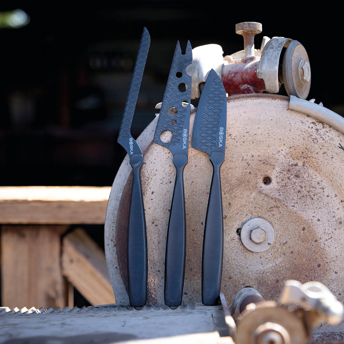 Three black cheese knives stand in front of a rusty saw and an old machine.