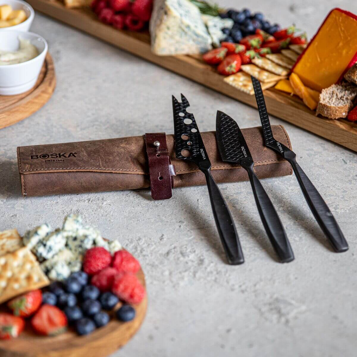 Black cheese knives rest on a leather roll next to cheese and fruit.