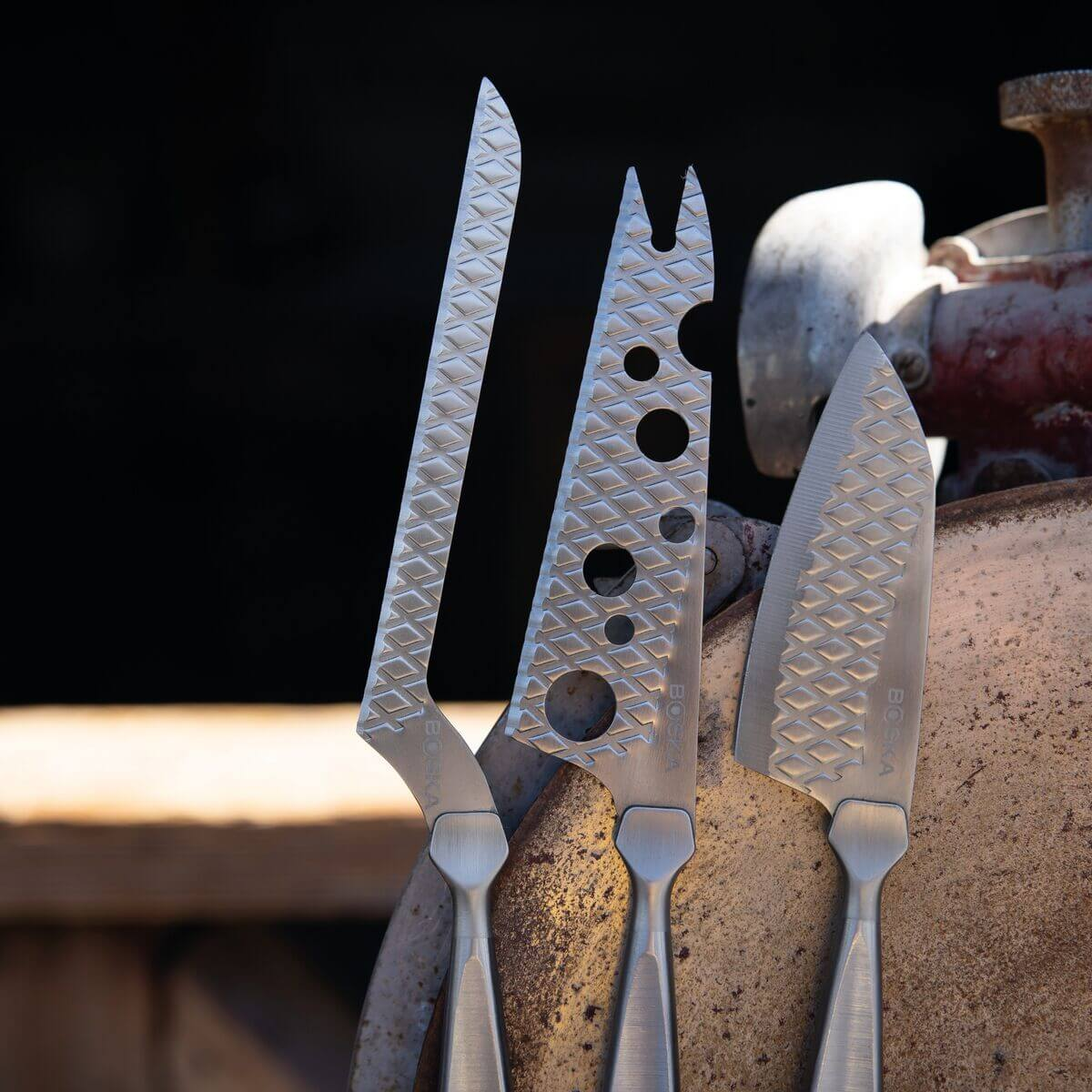 Three silver cheese knives with diamond pattern on a rusty surface.