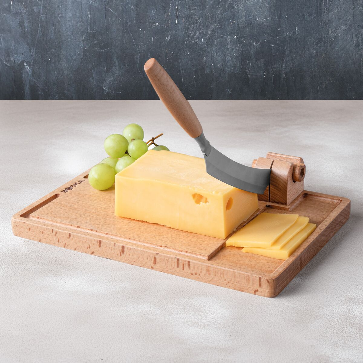 A block of cheese with grapes and a cheese knife on a wooden board.