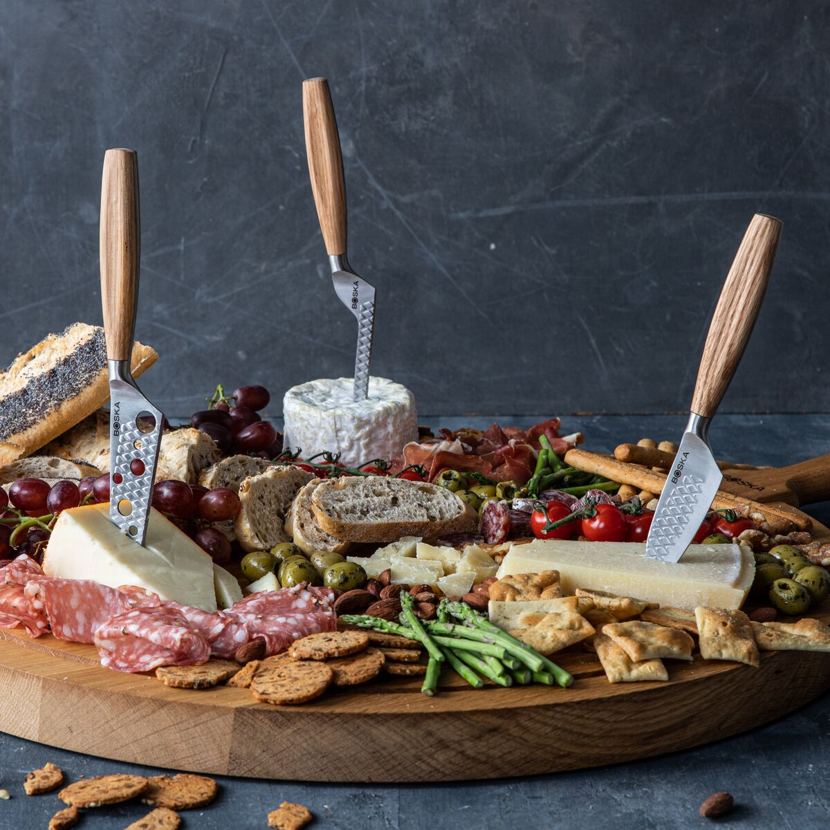 A large wooden board with a charcuterie spread: cheese, meats, olives, grapes, bread, and knives.