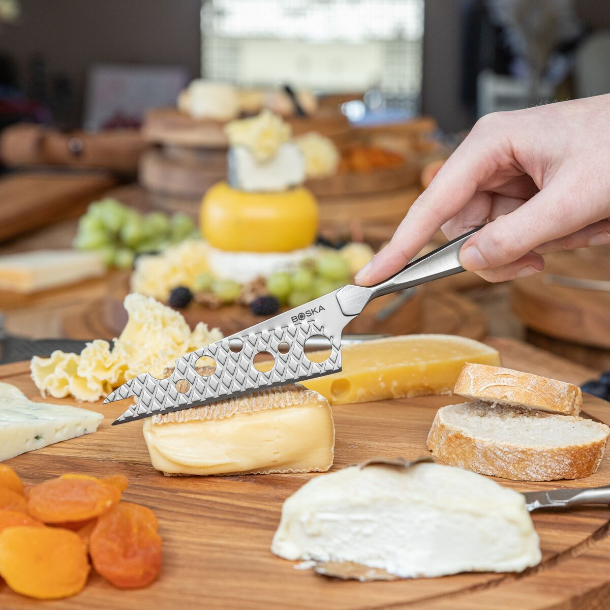 A hand uses a cheese knife to cut cheese on a wooden board with various cheeses.