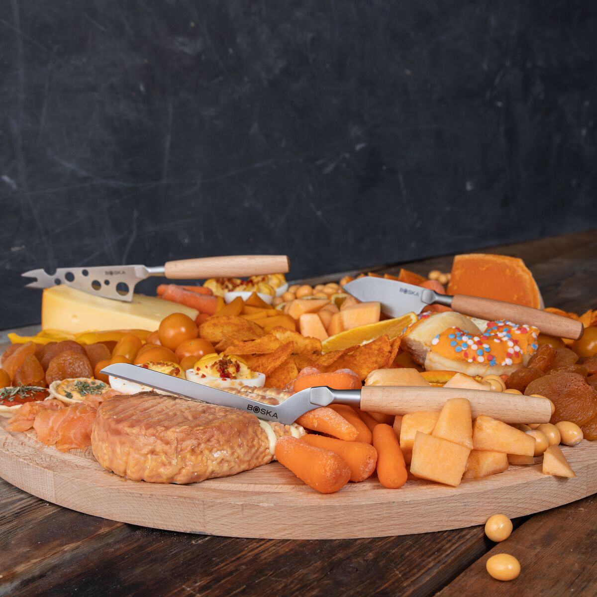A wooden board with cheese, meats, vegetables, fruits, and snacks, garnished with knives.