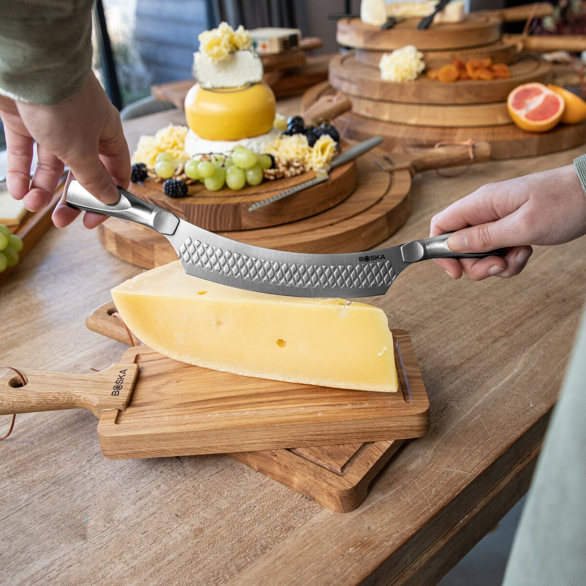 A person cuts a large block of cheese with a serrated knife on a wooden board.
