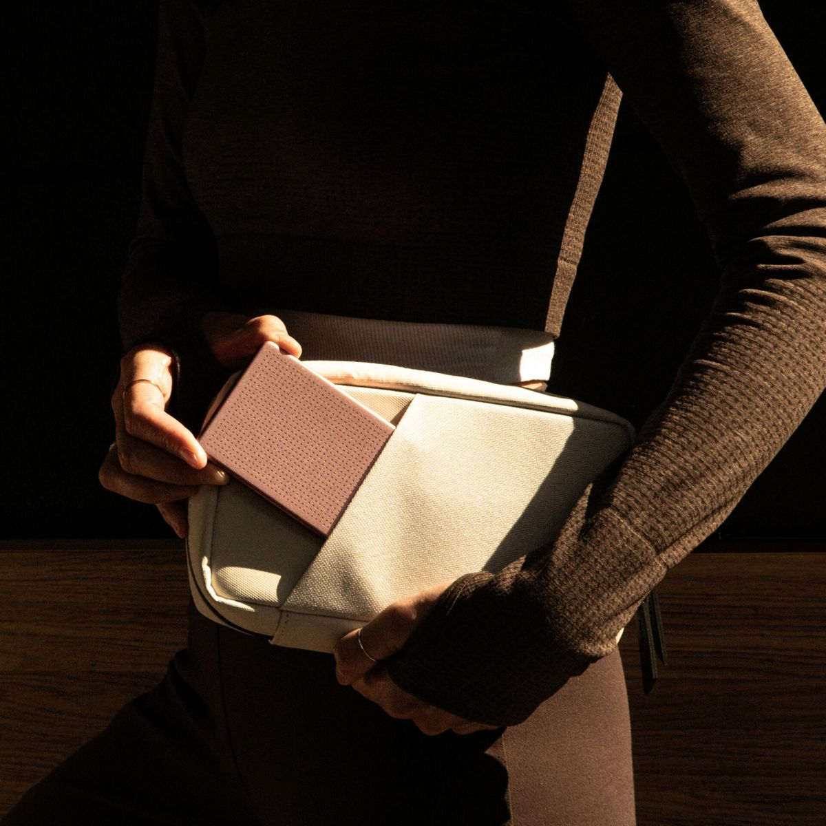 Una persona mette una scatola rettangolare rosa in una borsa a tracolla beige.