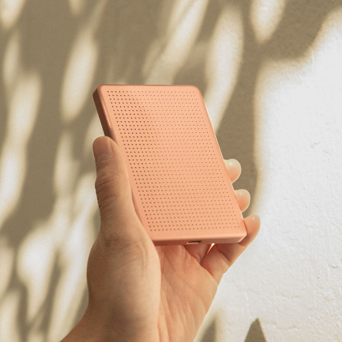 Una mano tiene un dispositivo rosa con motivo traforato. Ombre cadono sullo sfondo.