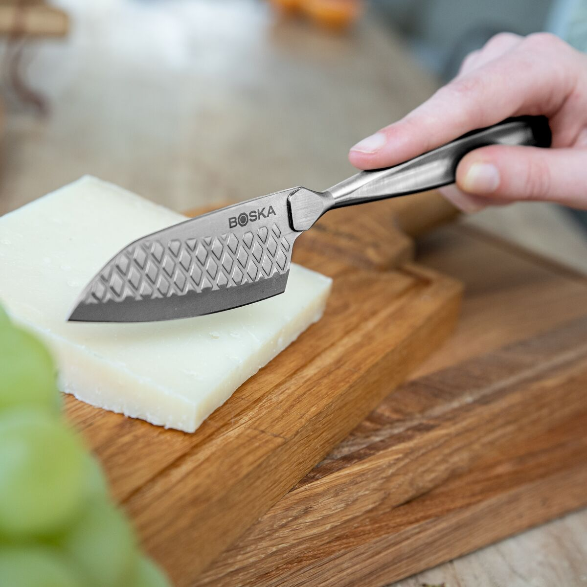 A knife cuts a piece of cheese on a wooden board, grapes in the foreground.