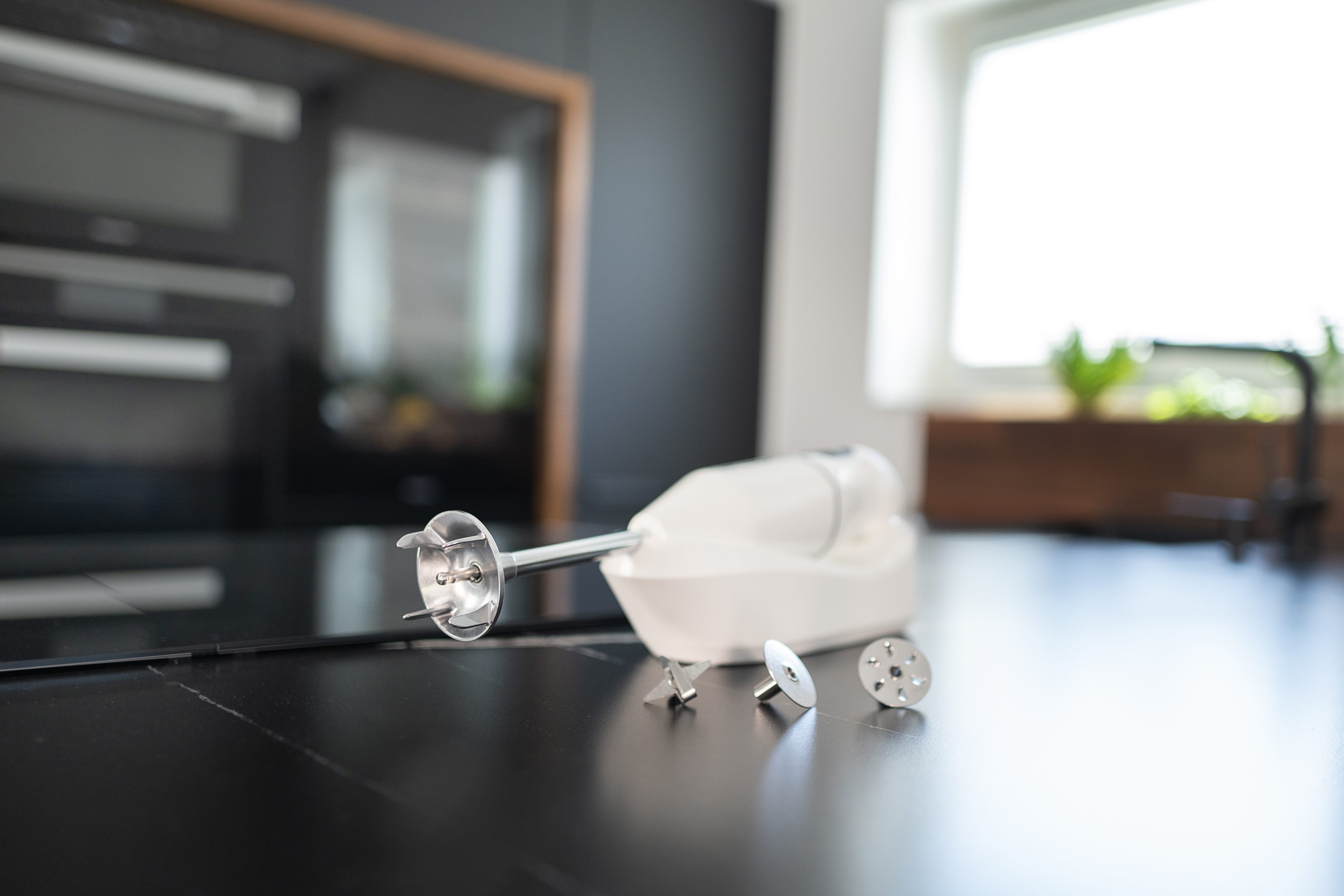 Biały blender ręczny z nasadkami spoczywa na ciemnym blacie.