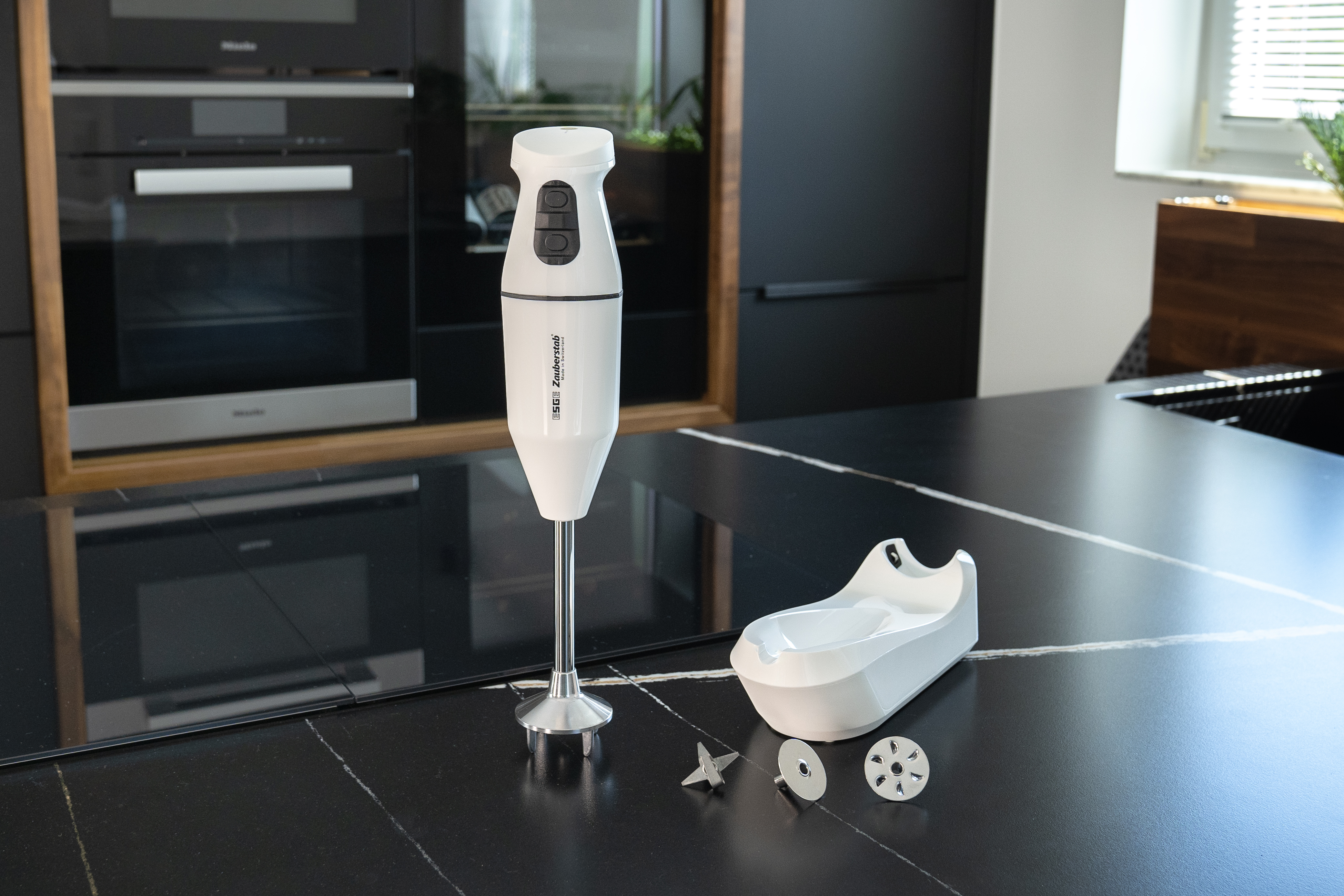 Biały blender ręczny z akcesoriami na czarnym stole przed sprzętem kuchennym.