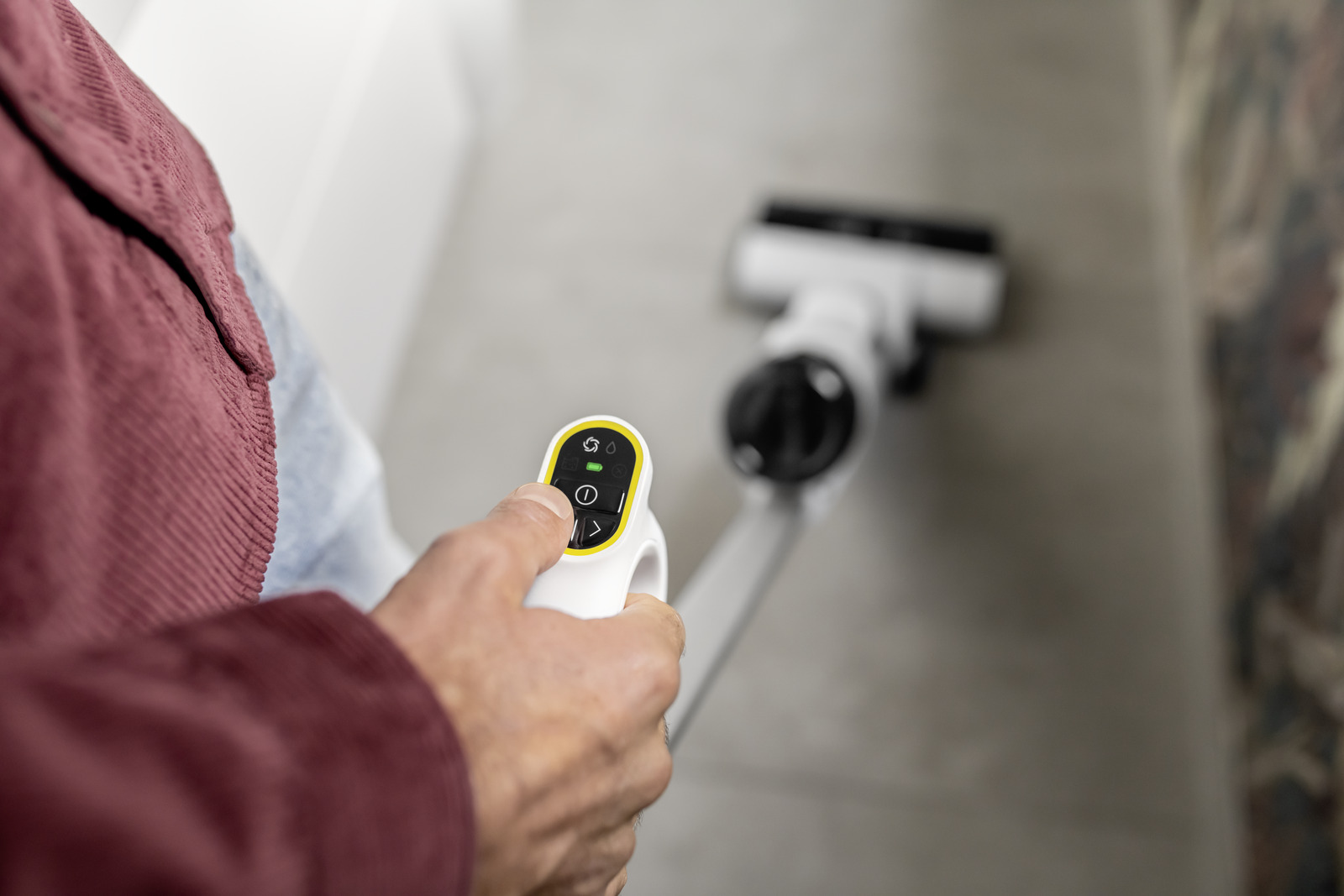 Ręka trzyma biały uchwyt odkurzacza z elementami sterującymi. Odkurzacz jest w tle.