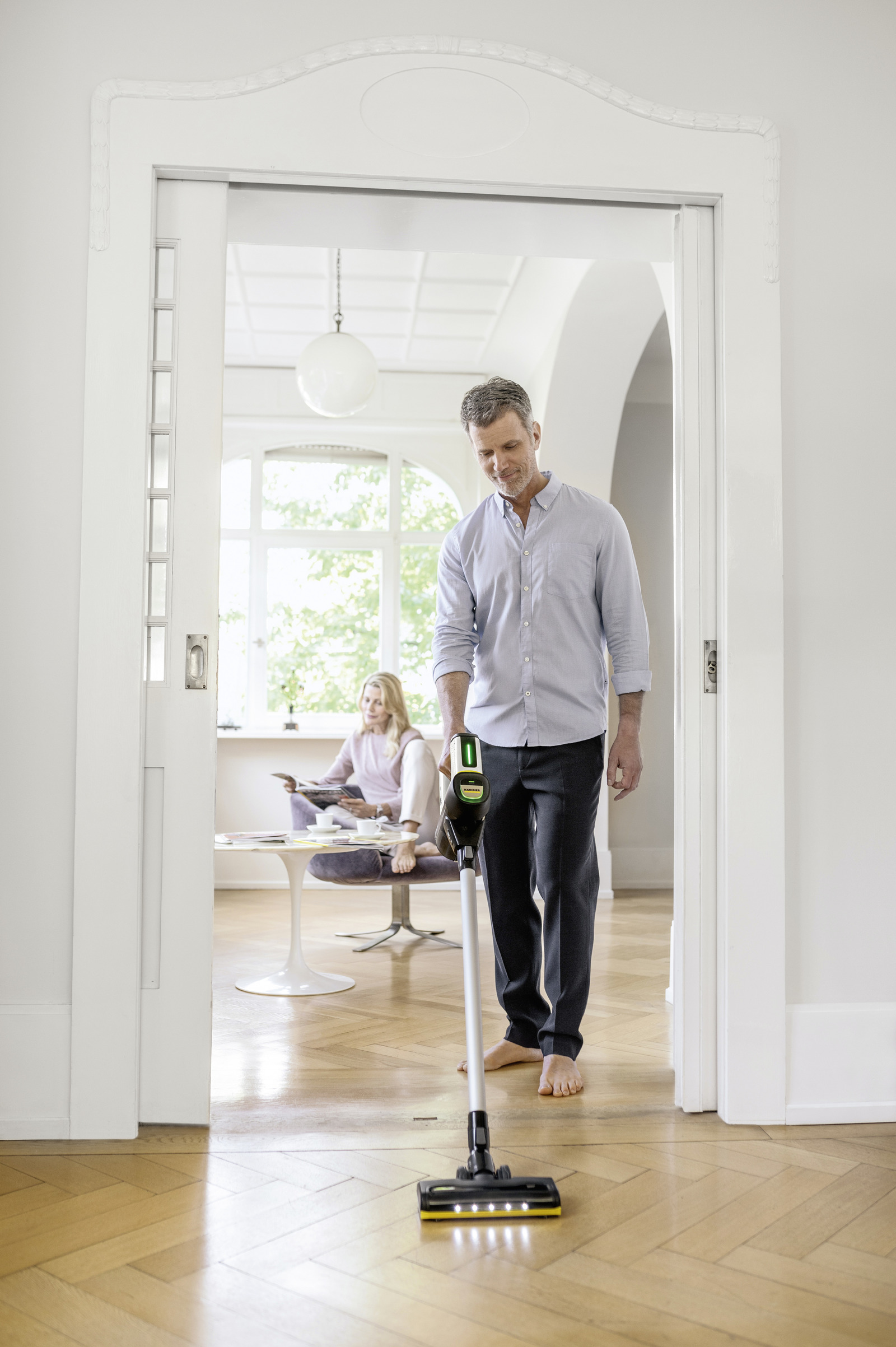 Homme passe l'aspirateur sur parquet, femme lit au fond.
