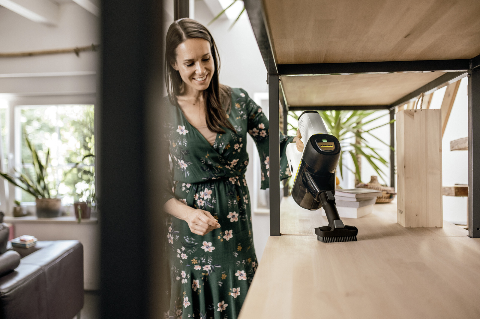 Kobieta czyści półkę odkurzaczem ręcznym, ubrana w zieloną sukienkę.