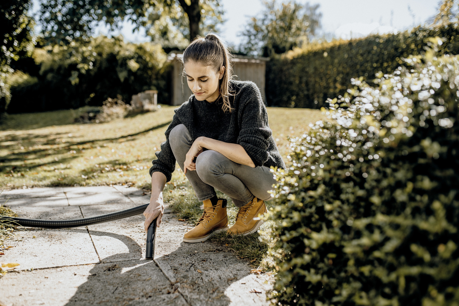 Kobieta z kucykiem czyści wężem wybrukowaną ścieżkę w ogrodzie.