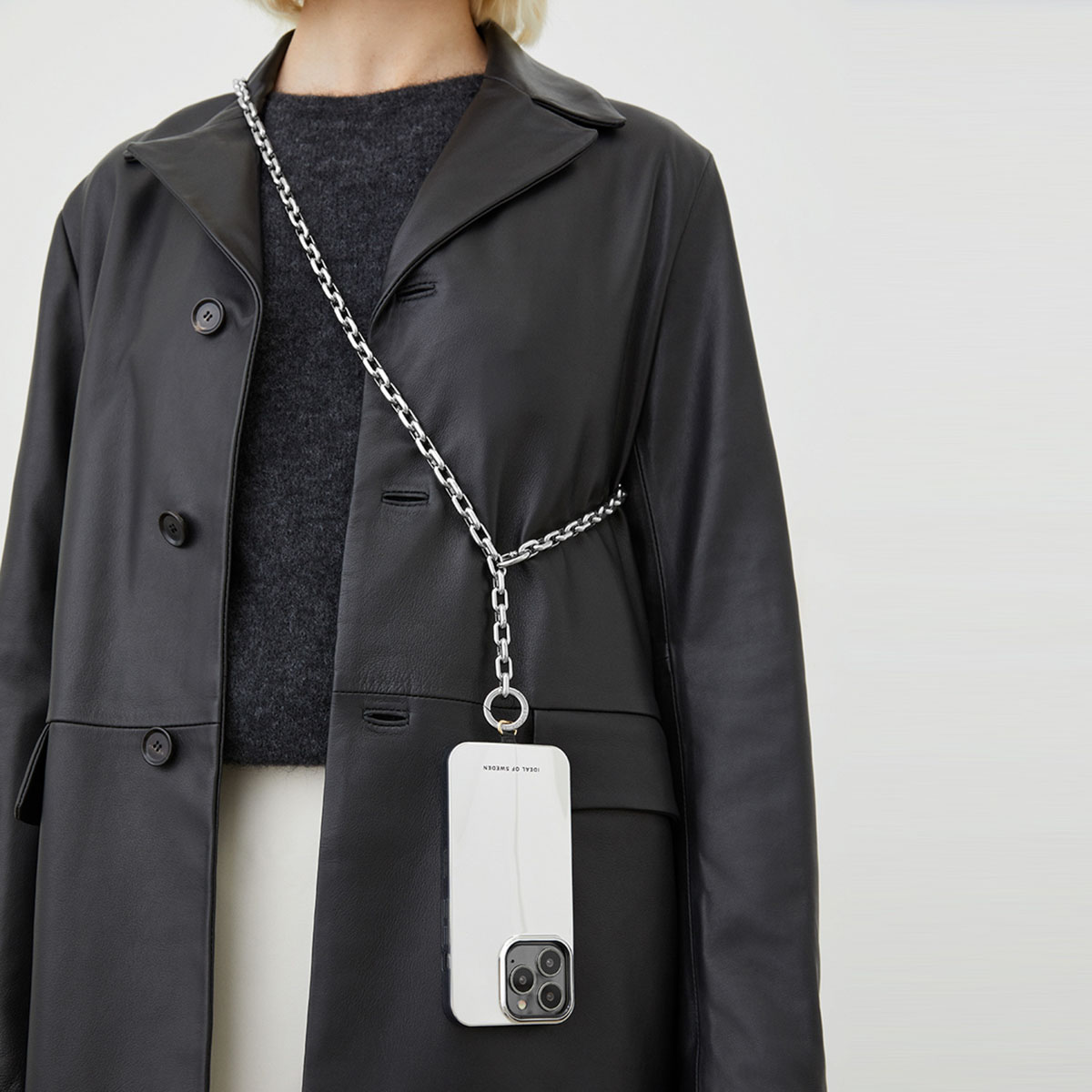 Eine Person trägt einen schwarzen Leder Trenchcoat und eine weiße Handyhülle mit Silberkette.
