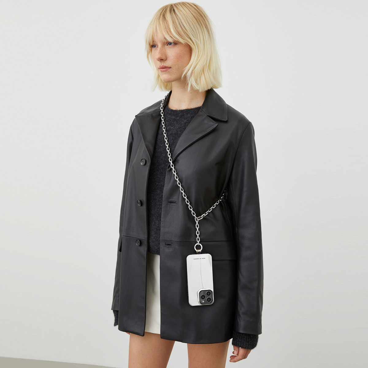 Eine Frau mit blondem Haar trägt eine schwarze Lederjacke, einen grauen Pullover und einen weißen Rock.