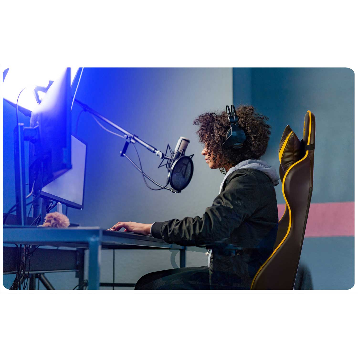 Kobieta w słuchawkach siedzi na fotelu gamingowym przed mikrofonem i komputerem.