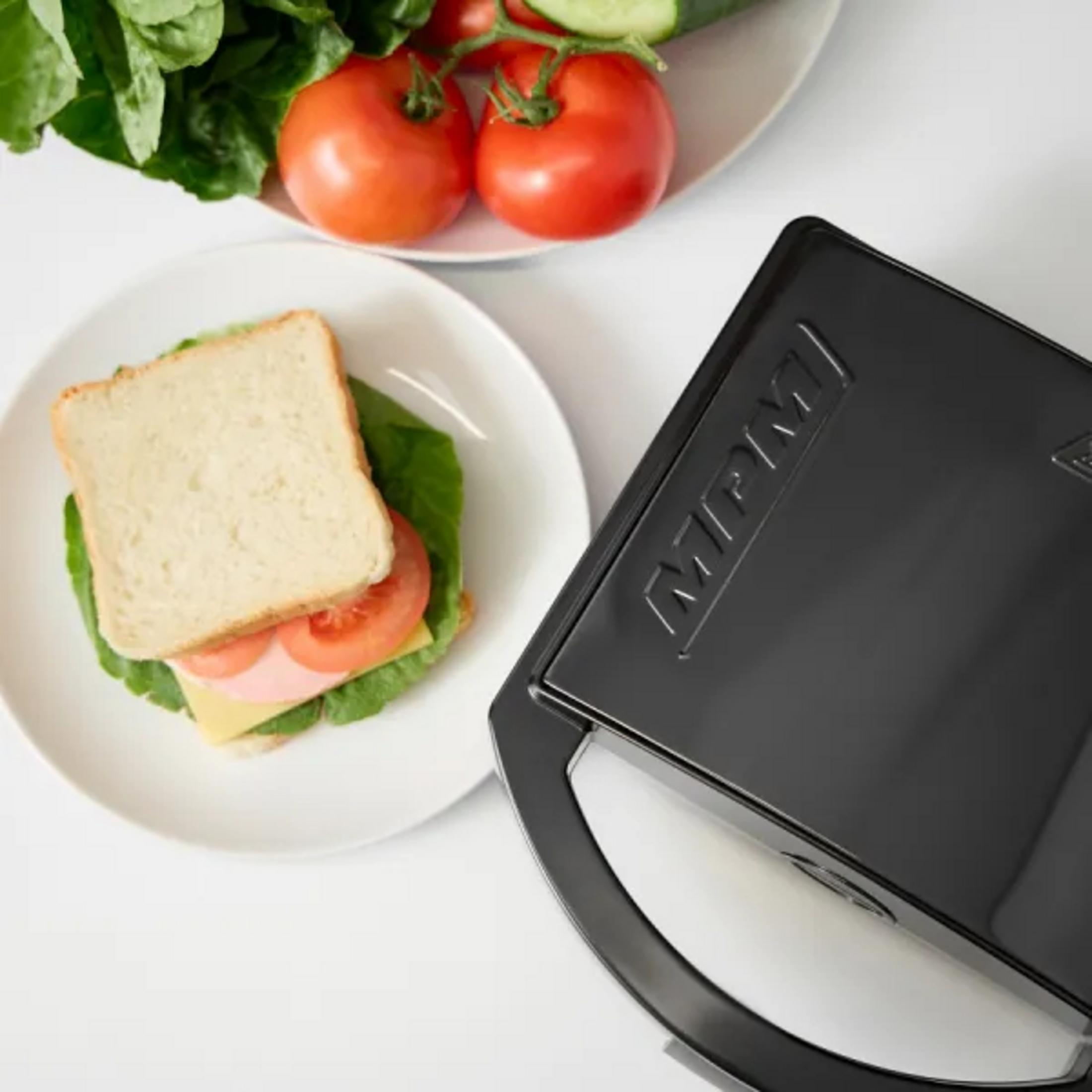 Un sandwich su un piatto accanto a una piastra per sandwich nera, pomodori e lattuga.