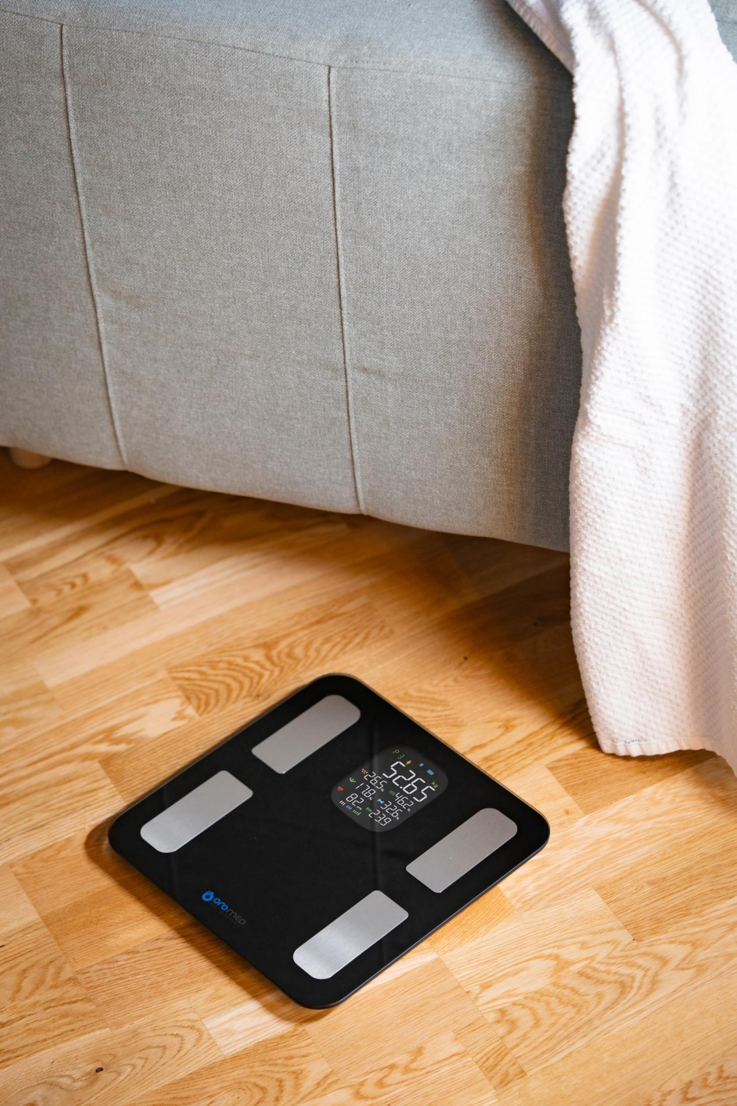 Bilancia digitale nera su pavimento in legno vicino a divano e asciugamano.