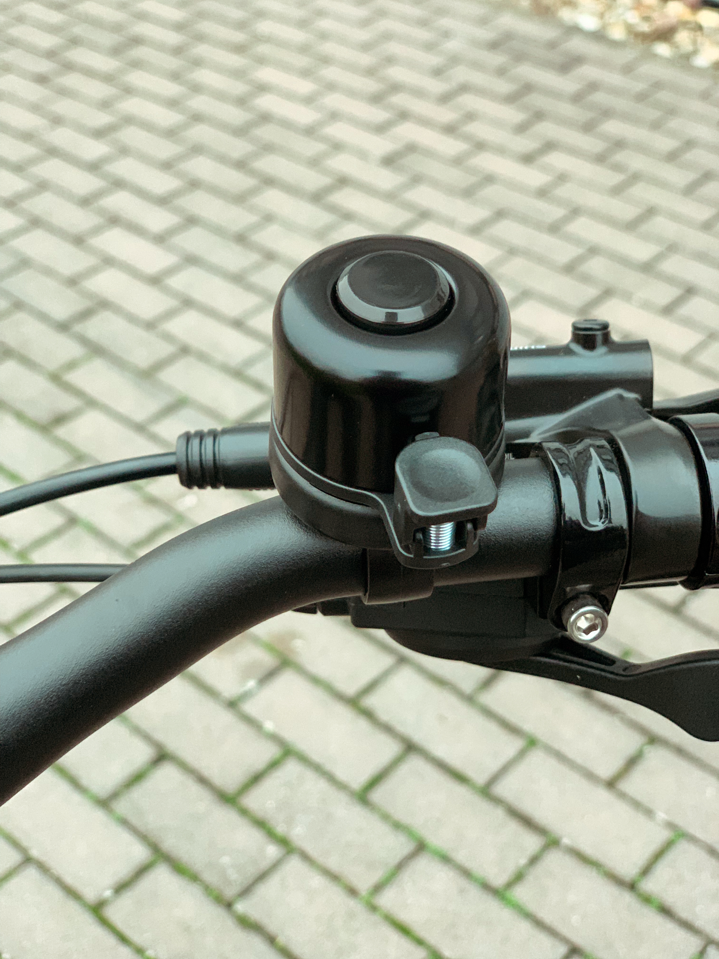 Schwarzer Fahrradklingel auf Lenker über gepflastertem Gehweg.