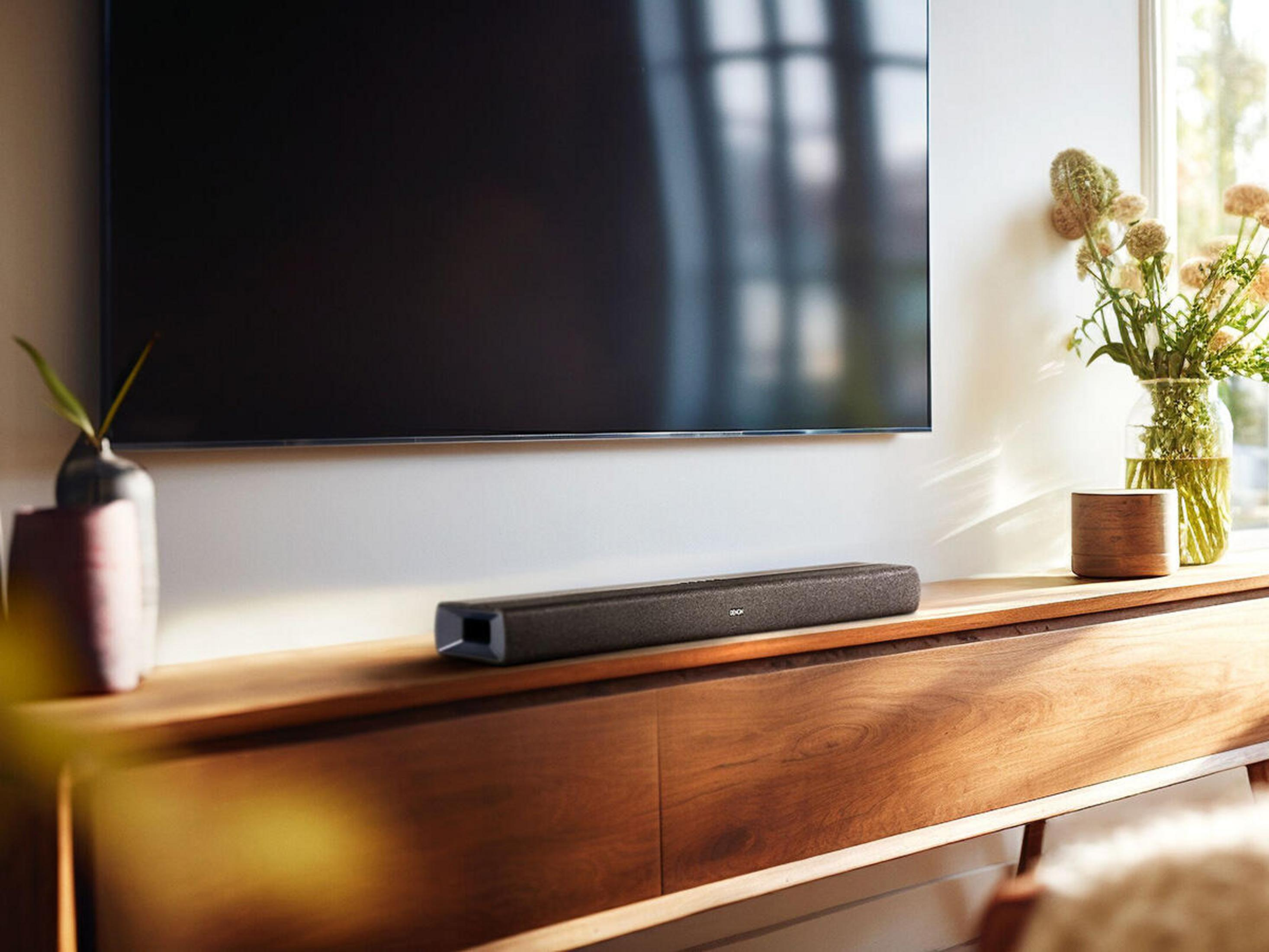 Ciemny soundbar leży na drewnym meblu pod ekranem telewizora.