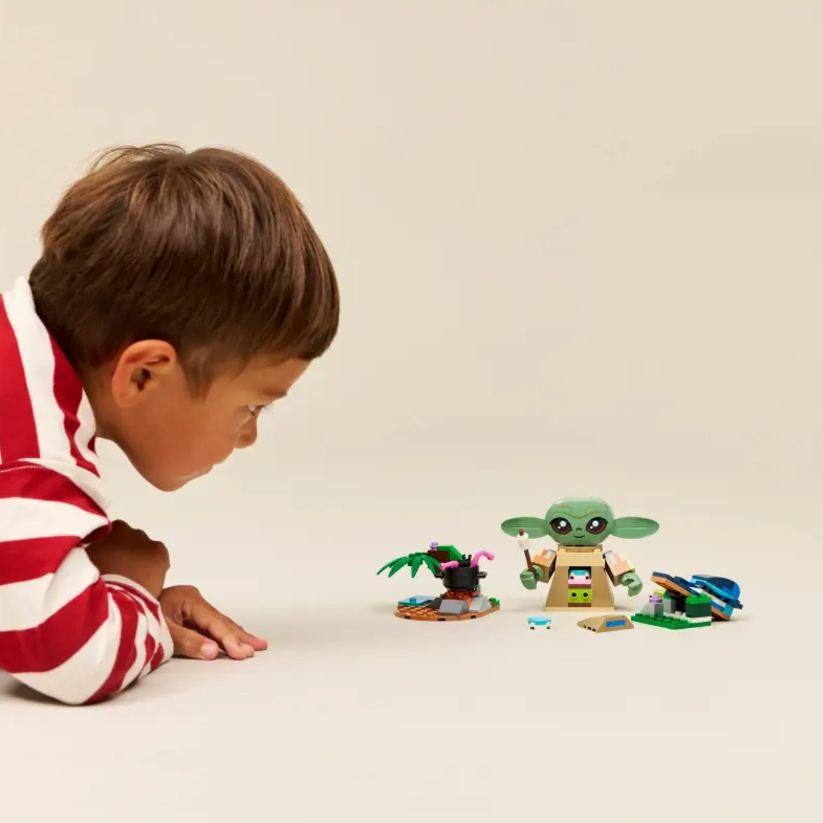Chłopiec w czerwono-białym swetrze patrzy na małą zieloną figurkę i zabawkowe otoczenie.