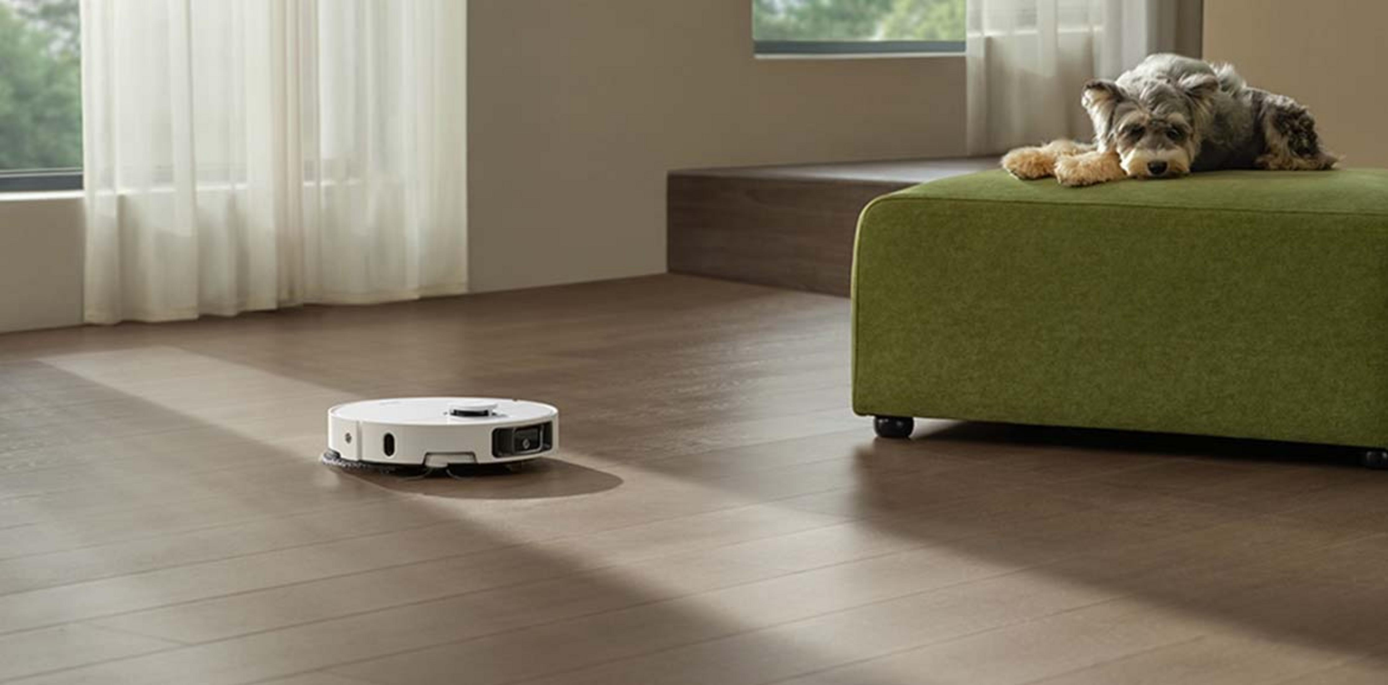 Un robot aspirapolvere bianco pulisce un pavimento in legno mentre un cane riposa su un pouf verde.