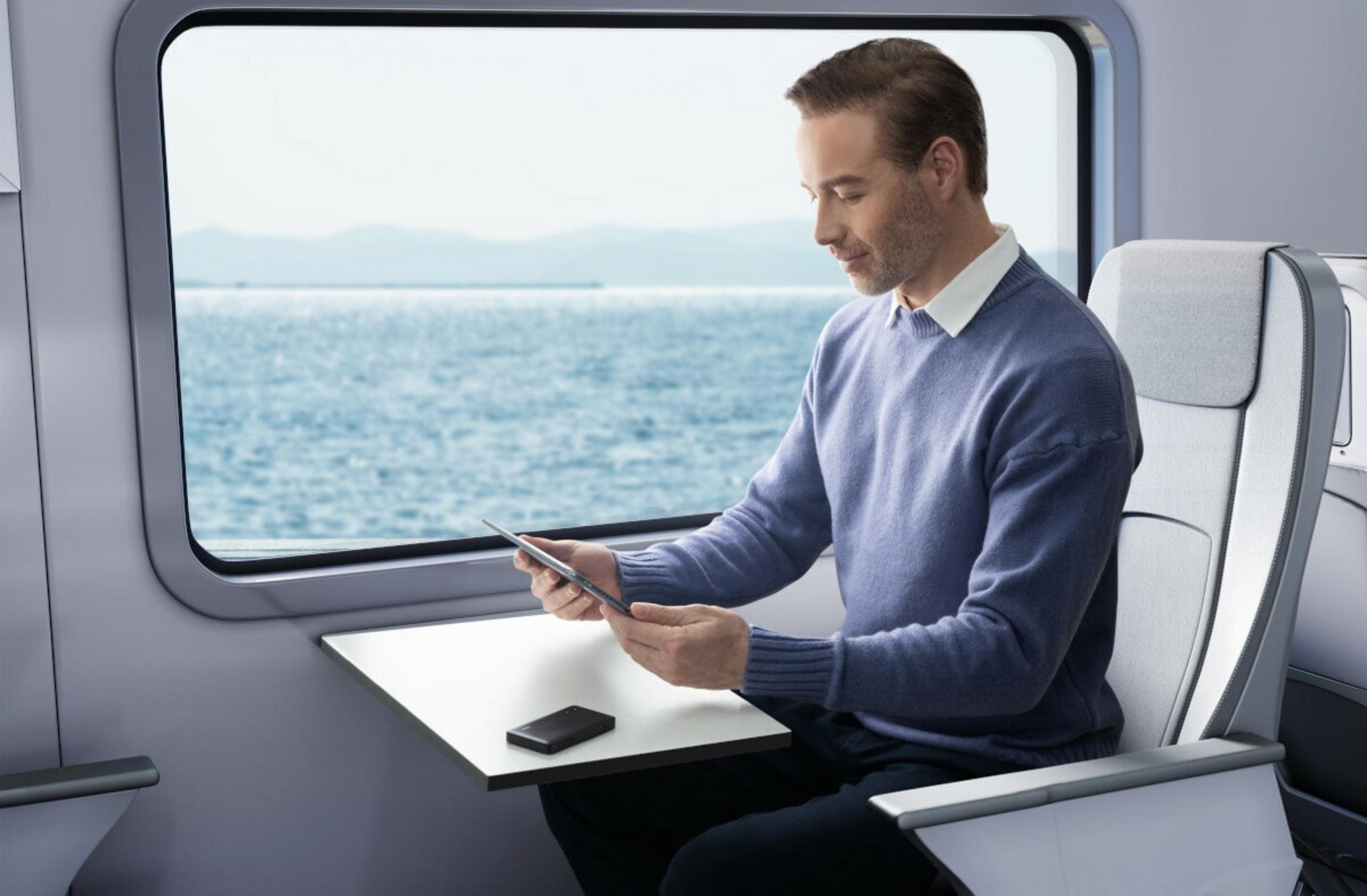 Uomo in maglione blu con tablet su treno affacciato sul mare.