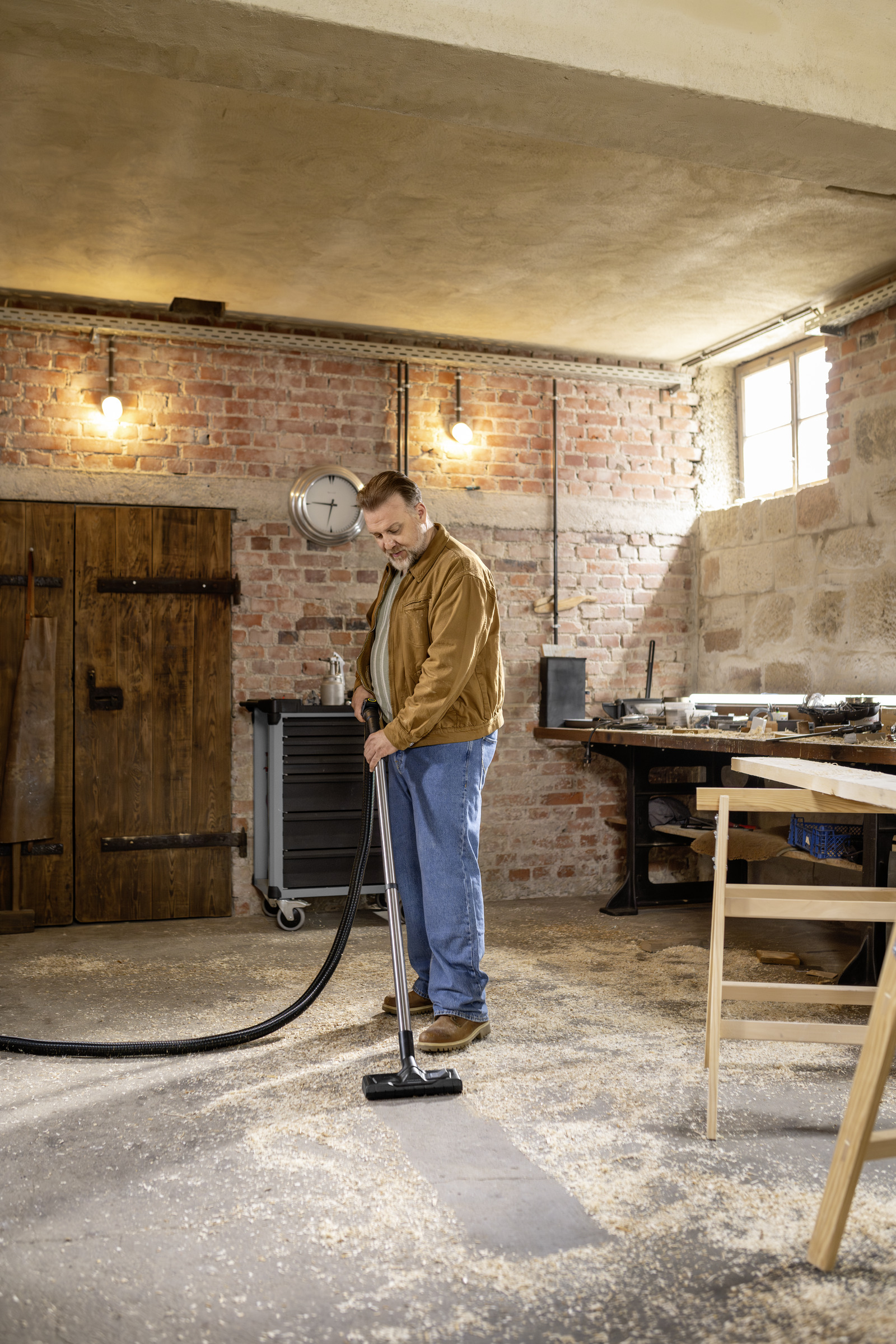 A man vacuums wood shavings in a workshop. A wooden door and workbenches are in the background.