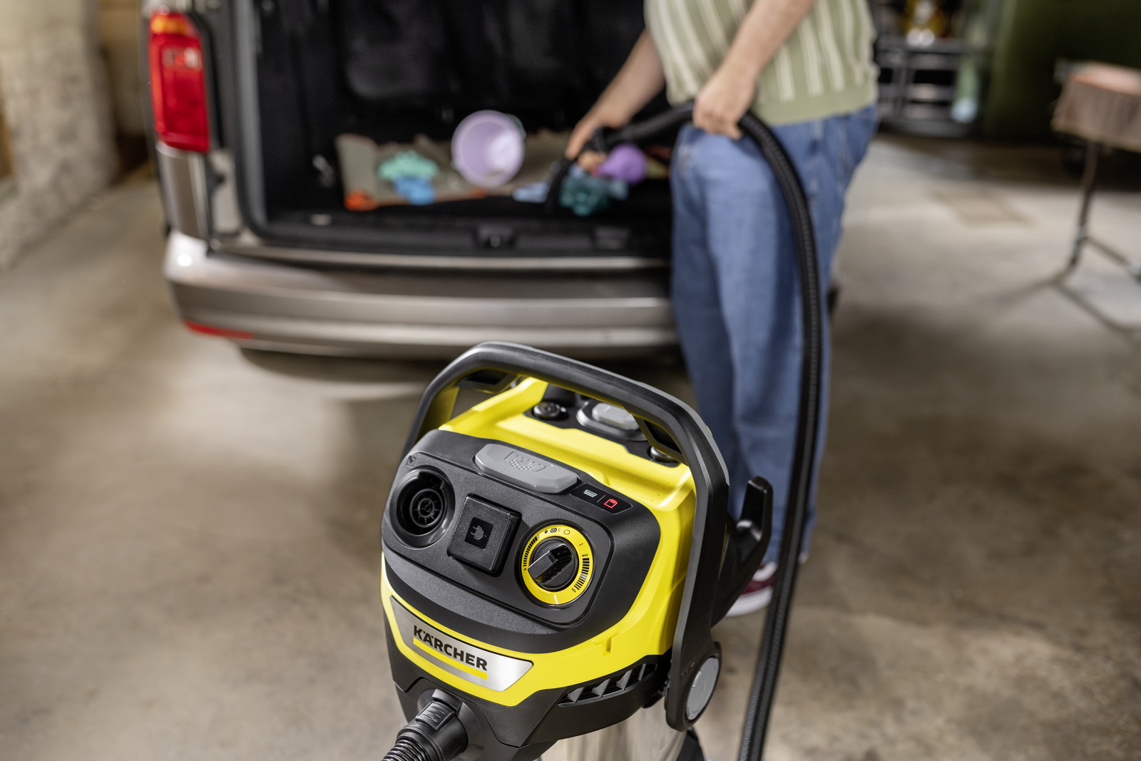 Yellow wet/dry vacuum in foreground, person vacuuming car trunk in background.