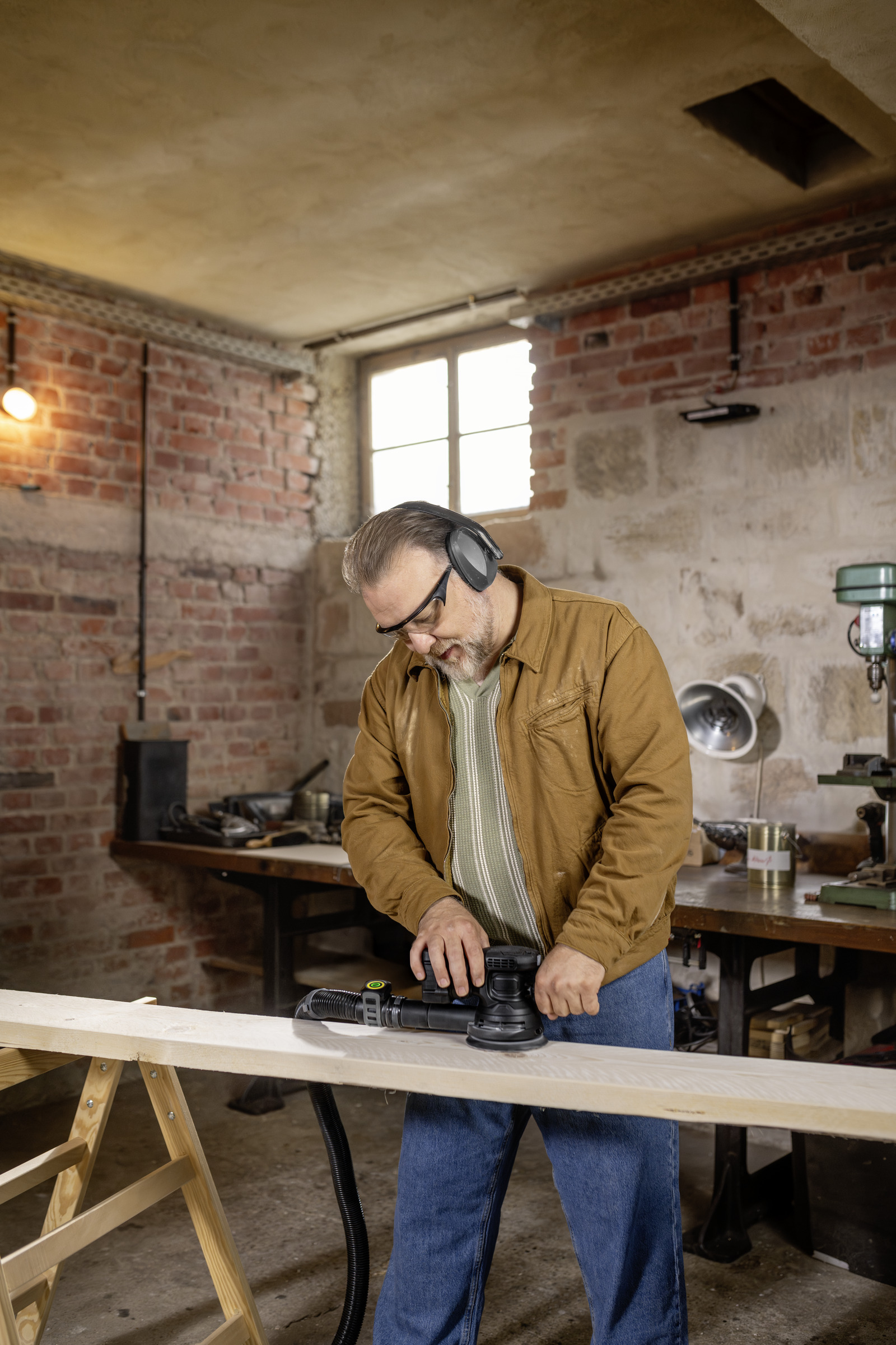 Man sanding a wooden board in a workshop with a sander and safety glasses.