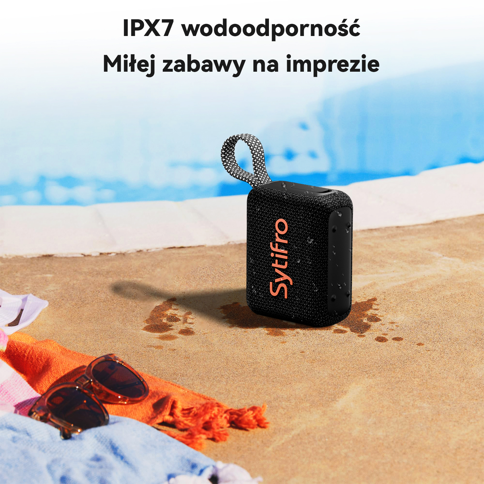 Czarny głośnik z pomarańczowym logo na piaszczystym brzegu basenu, okulary przeciwsłoneczne i ręczniki.