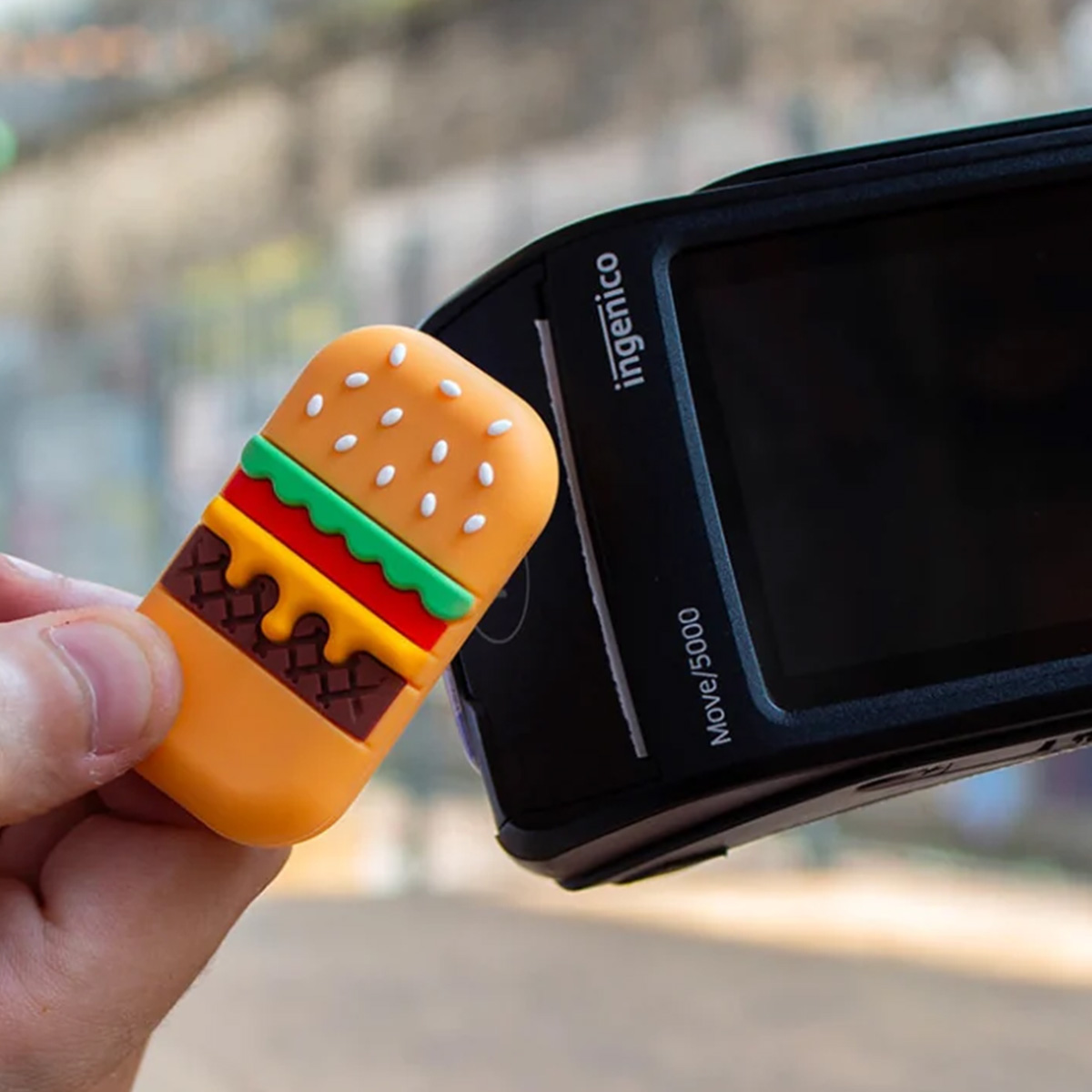 Un'unità USB a forma di hamburger è tenuta accanto a un terminale di pagamento Ingenico Move/5000 nero.