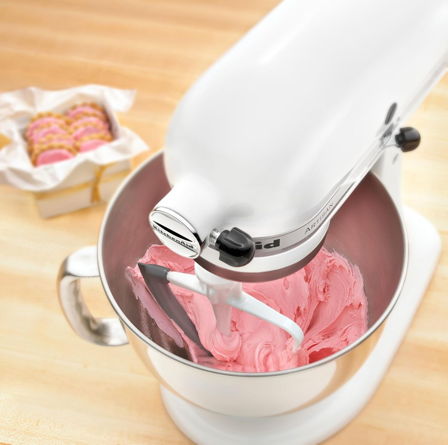 Eine weiße Küchenmaschine mit rosa Zuckerguss in einer Schüssel und Keksen im Hintergrund.