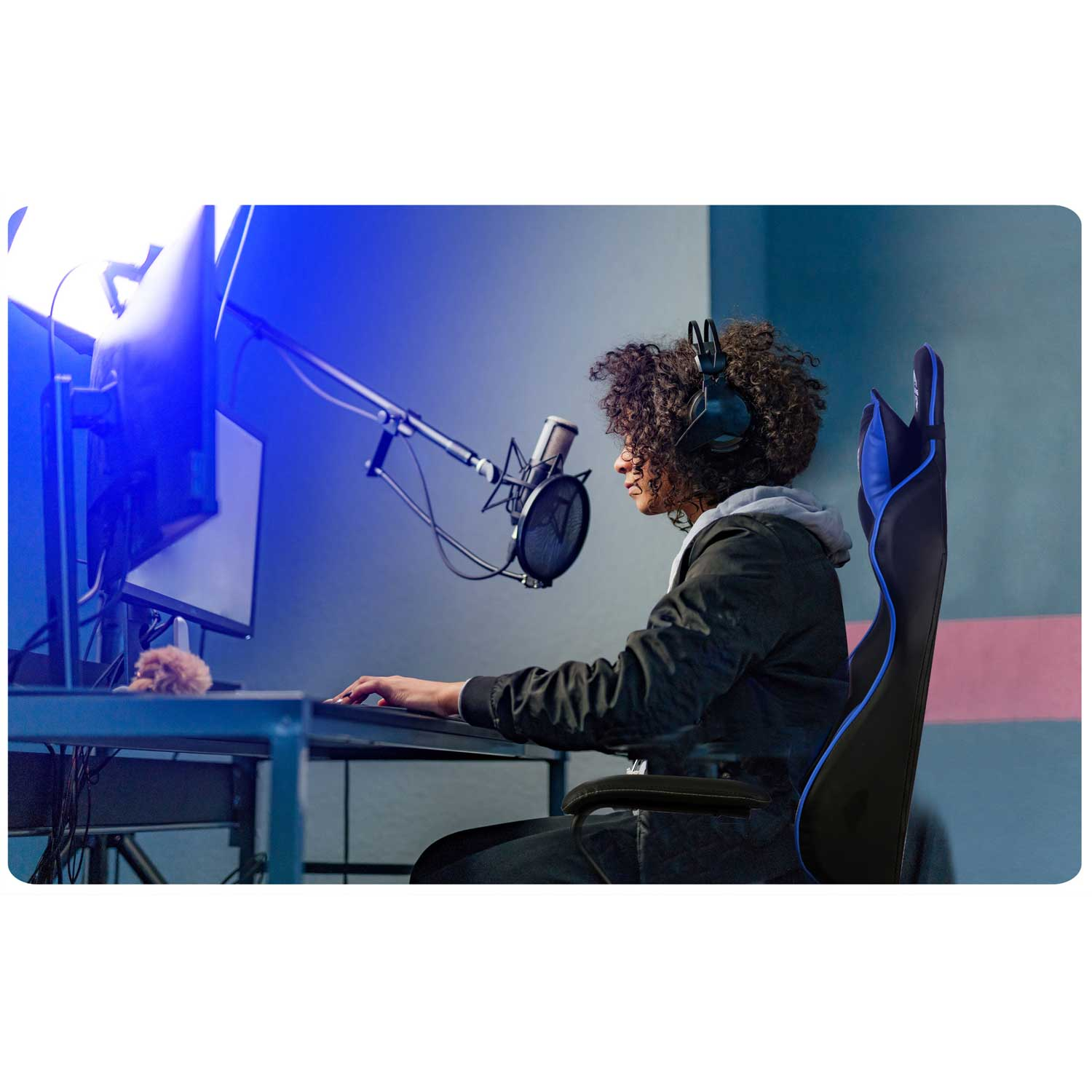 Kobieta w słuchawkach siedzi w fotelu gamingowym przed mikrofonem i dwoma monitorami.