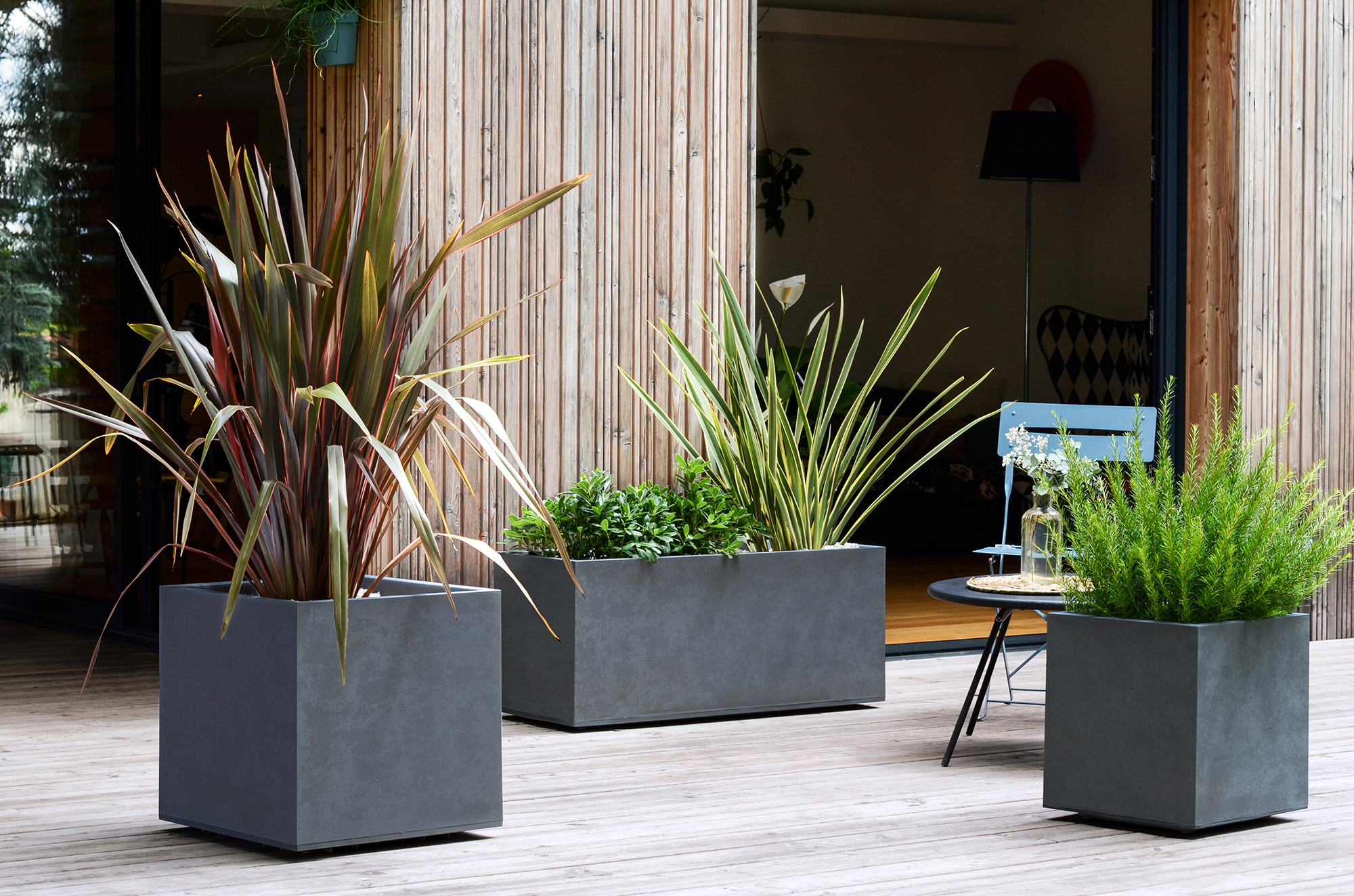 Moderne Betonpflanzkübel mit grünen Pflanzen auf einer Holzterrasse.