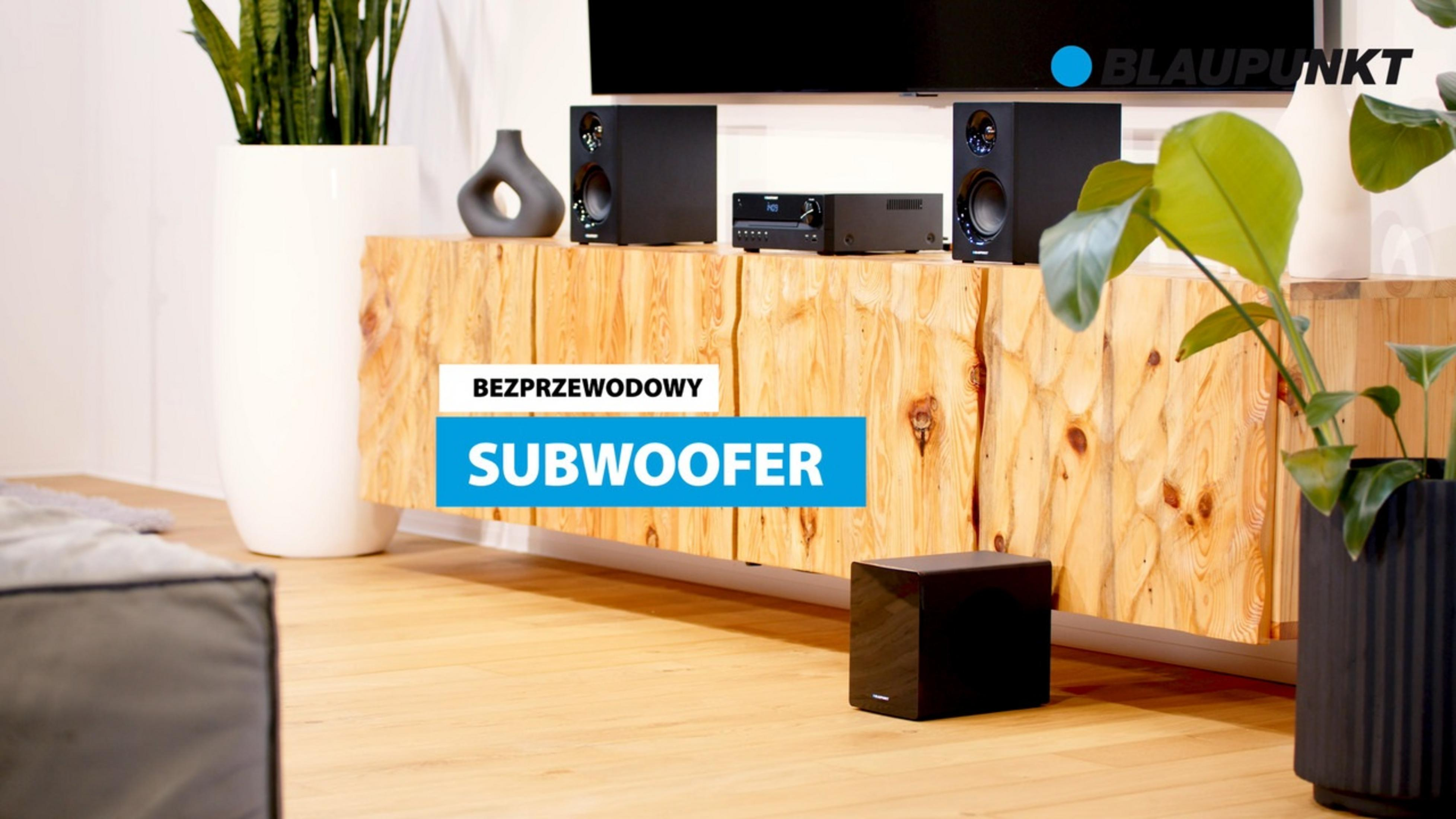 Un subwoofer nero siede su un pavimento in legno davanti a un mobile in legno con altoparlanti.